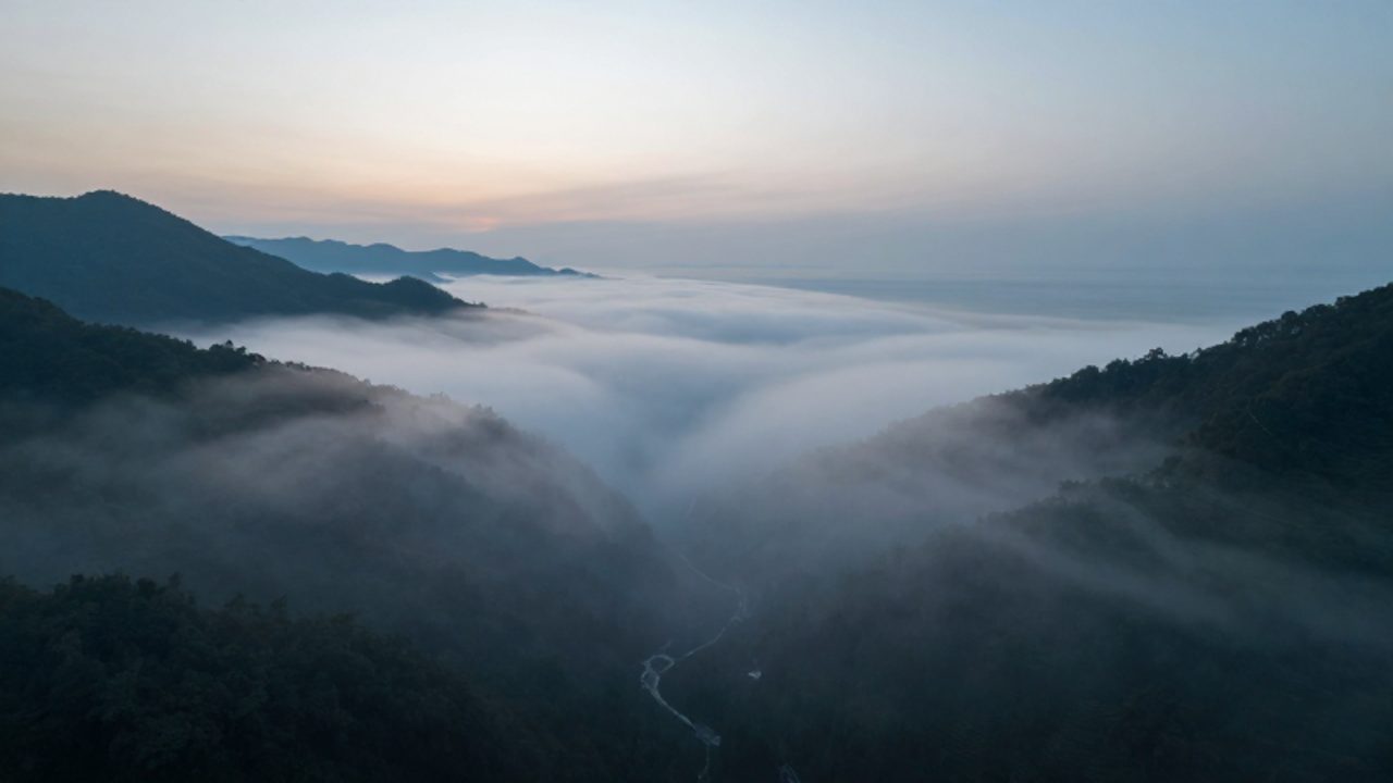Paisaje de montaña y costa donde se observa niebla densa acumulada en un valle y bancos de niebla desplazándose sobre la línea de costa al amanecer.