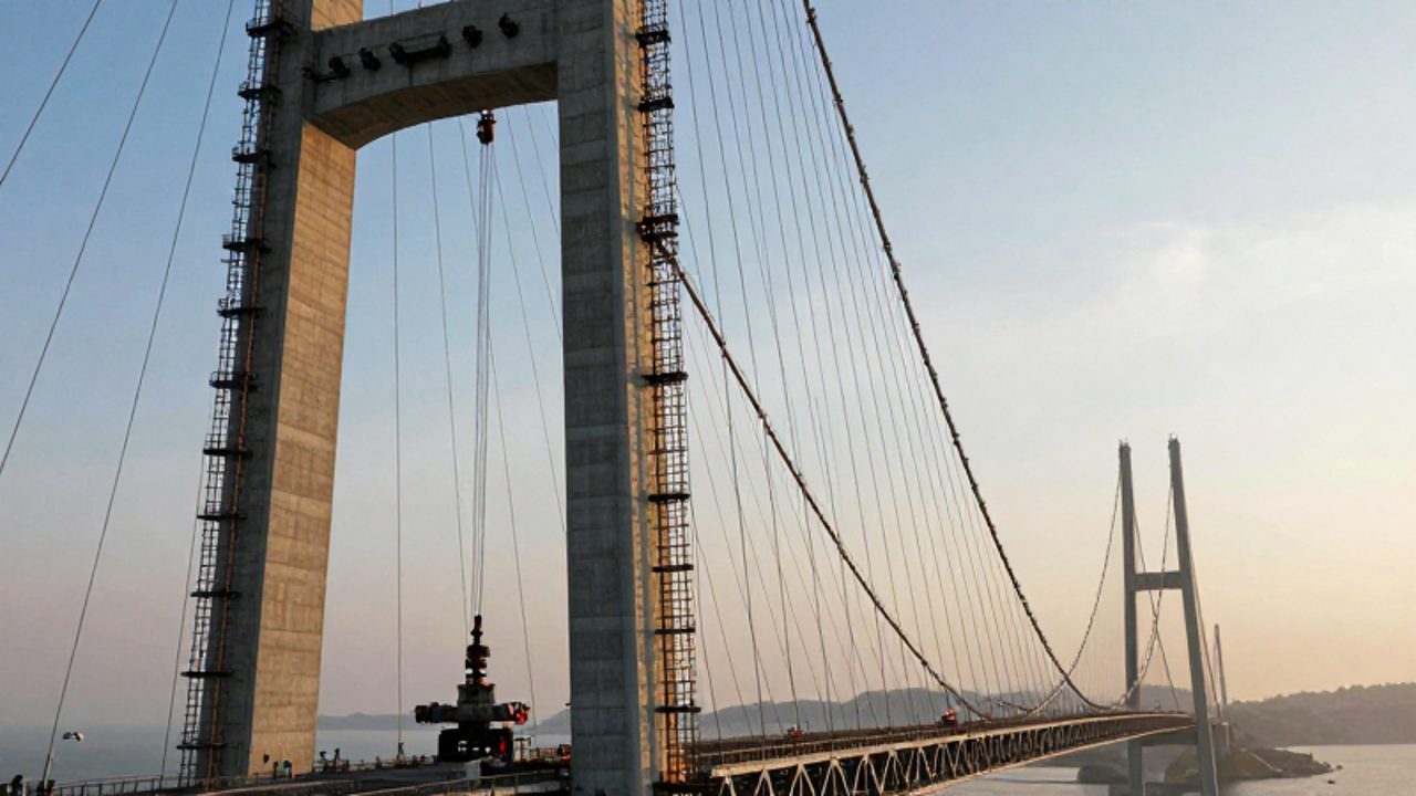 Torres de hormigón y una red de cables de acero de un puente colgante en construcción sobre un estrecho marino durante el atardecer.