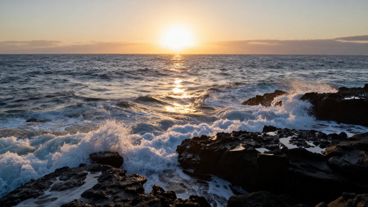 El sol se alza bajo en el horizonte proyectando un reflejo dorado sobre las olas que rompen contra rocas volcánicas en la costa.