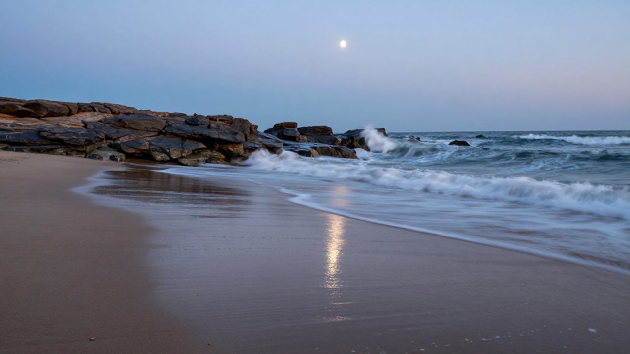 Costa rocosa al atardecer donde se aprecia la transición entre la marea baja y el avance del océano bajo la luz de una luna creciente en el horizonte.