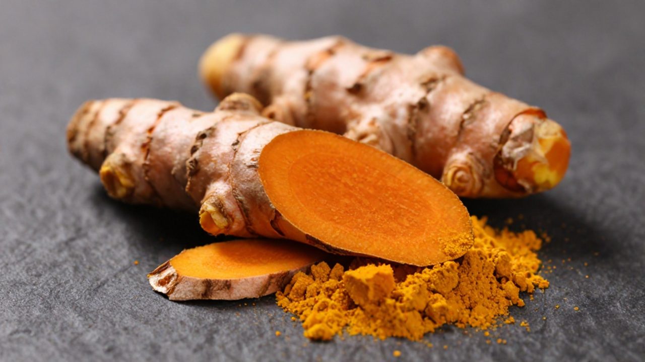 Raíces de cúrcuma partidas que muestran su interior naranja intenso sobre una superficie oscura rodeada de polvo de la misma especia.