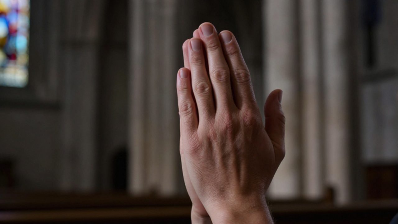 Manos unidas en gesto de oración frente a la arquitectura tenue de una catedral antigua.